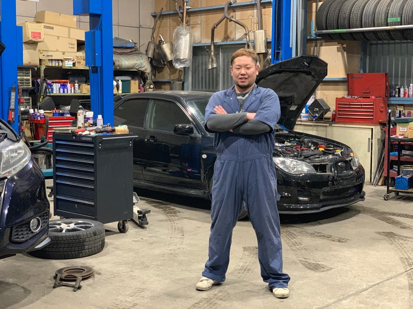 Sato Tasuhiko in overalls in the workshop, in front of a car with its bonnet open